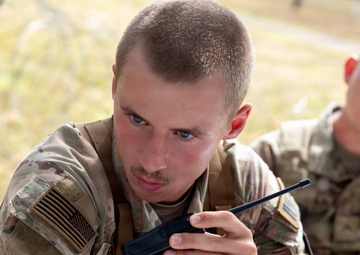 Two U.S. Army soldiers in Operational Camouflage Pattern (OCP) uniforms are engaged in what appears to be a field training exercise or operational task. The foreground soldier is holding a handheld radio/walkie-talkie and appears focused on a task, while a second soldier wearing a patrol cap observes from behind. Documents or notebooks are visible on a surface in front of them.