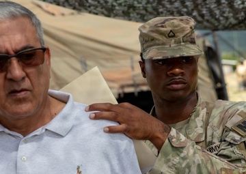 A U.S. Army Military Police (MP) soldier in OCP camouflage uniform has his hand placed on the shoulder of a civilian man wearing a white polo shirt. The setting appears to be a military field exercise or base, with tactical tents, camouflage netting, and military vehicles visible in the background. The interaction appears to be part of a training scenario, possibly simulating detainee handling or civilian control procedures.
