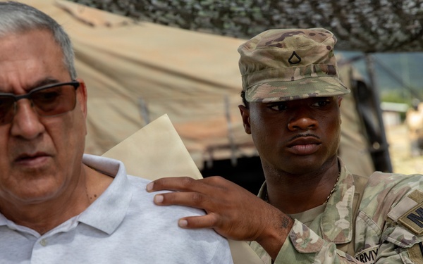A U.S. Army Military Police (MP) soldier in OCP camouflage uniform has his hand placed on the shoulder of a civilian man wearing a white polo shirt. The setting appears to be a military field exercise or base, with tactical tents, camouflage netting, and military vehicles visible in the background. The interaction appears to be part of a training scenario, possibly simulating detainee handling or civilian control procedures.