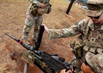 U.S. Army Military Police (MP) soldiers are conducting weapons training with what appears to be an M240 machine gun mounted on a tripod. Two soldiers in the foreground are actively handling and inspecting the weapon, while a third soldier stands in the background observing. All personnel are wearing Operational Camouflage Pattern (OCP) uniforms on what appears to be a military training range.