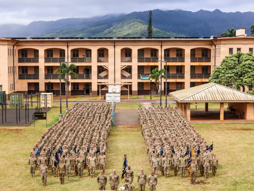A large military formation is assembled on a parade ground in front of a multi-story tan/salmon-colored barracks building. Hundreds of soldiers in camouflage uniforms are arranged in two large rectangular formations flanking a central pathway, with flag bearers and officers positioned at the front. The setting appears to be a tropical military installation, likely in Hawaii, evidenced by the lush green mountains, palm trees, and architectural style in the background.