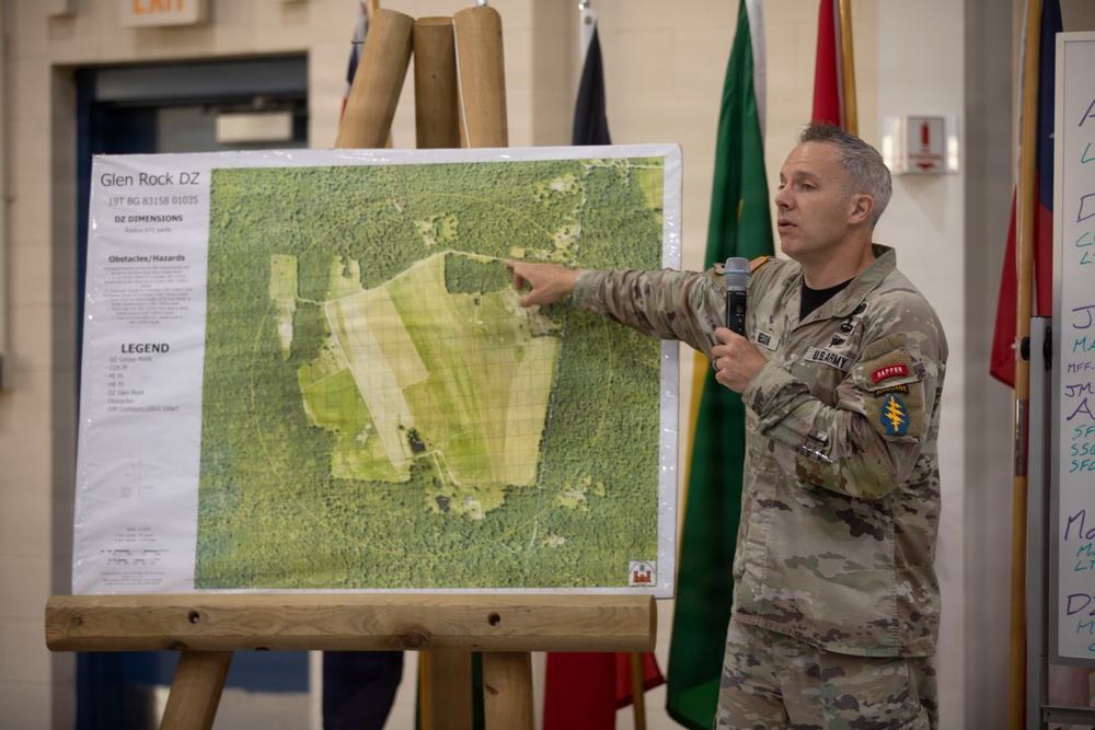 A U.S. Army soldier in MultiCam/OCP camouflage uniform is conducting a briefing, pointing to a large aerial map mounted on a wooden easel. The map is labeled 'Glen Rock DZ' (Drop Zone) with coordinates '19T BG 83158 01035' and includes DZ dimensions, obstacles/hazards, and a legend. The setting appears to be an indoor military facility with unit flags visible in the background.
