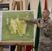 A U.S. Army soldier in MultiCam/OCP camouflage uniform is conducting a briefing, pointing to a large aerial map mounted on a wooden easel. The map is labeled 'Glen Rock DZ' (Drop Zone) with coordinates '19T BG 83158 01035' and includes DZ dimensions, obstacles/hazards, and a legend. The setting appears to be an indoor military facility with unit flags visible in the background.