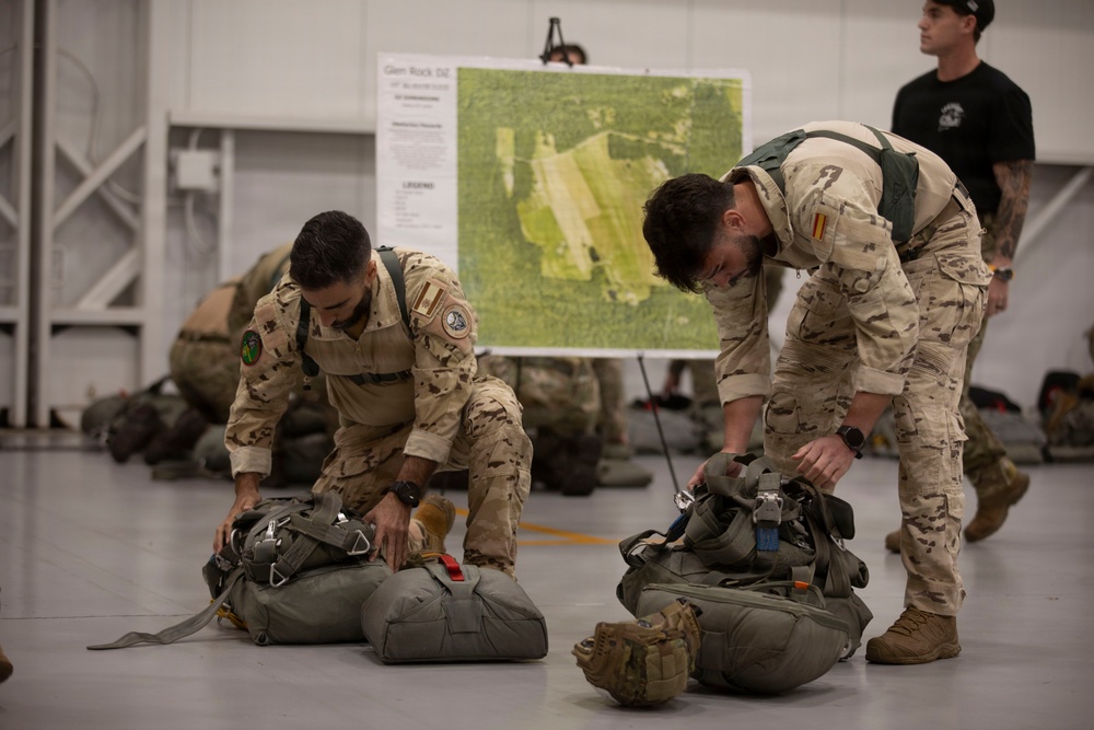 Two soldiers in desert camouflage uniforms are inspecting or packing parachute equipment on the floor of what appears to be a military hangar. A aerial map labeled 'Glen Rock DZ' (Drop Zone) is visible on a board in the background, suggesting pre-jump mission planning. A third individual in a black t-shirt observes from the background.