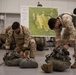 Two soldiers in desert camouflage uniforms are inspecting or packing parachute equipment on the floor of what appears to be a military hangar. A aerial map labeled 'Glen Rock DZ' (Drop Zone) is visible on a board in the background, suggesting pre-jump mission planning. A third individual in a black t-shirt observes from the background.