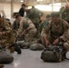 Multiple military personnel from different nations are gathered inside a large hangar, preparing parachute equipment on the floor. Soldiers wearing various national uniforms, including Italian and British camouflage patterns, are kneeling and checking their gear. The scene appears to be a pre-jump equipment inspection or rigging exercise during a multinational airborne training operation.
