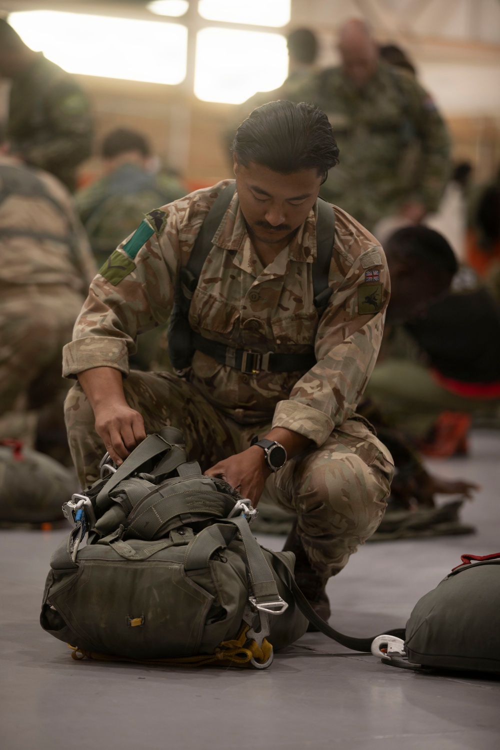 A British Army soldier in Multi-Terrain Pattern (MTP) camouflage uniform is kneeling on a hangar floor, inspecting or adjusting parachute equipment. He is wearing a parachute harness and is focused on a packed parachute container in front of him. Multiple other soldiers in the background appear to be conducting similar pre-jump equipment checks.