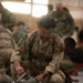 A British Army soldier in Multi-Terrain Pattern (MTP) camouflage uniform is kneeling on a hangar floor, inspecting or adjusting parachute equipment. He is wearing a parachute harness and is focused on a packed parachute container in front of him. Multiple other soldiers in the background appear to be conducting similar pre-jump equipment checks.