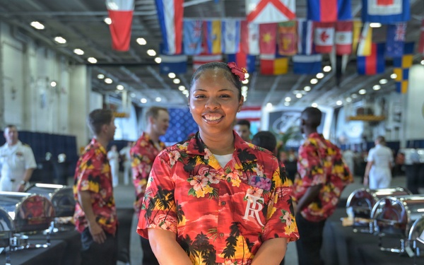A smiling food service worker wearing a red Hawaiian/tropical print shirt and blue latex gloves stands behind a serving station in what appears to be a large military vessel hangar bay. The space is decorated with numerous international flags hanging from the ceiling, suggesting a special event or celebration. Multiple chafing dishes with heat lamps are set up along serving tables, indicating a buffet-style meal service.