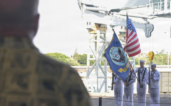 A U.S. Navy color guard ceremony is taking place on what appears to be the deck of a naval vessel or pier facility. Three sailors in dress white uniforms are presenting the American flag and a blue unit/command flag while armed with rifles. A civilian in a Hawaiian/aloha shirt and a military officer observe the ceremony from the foreground.