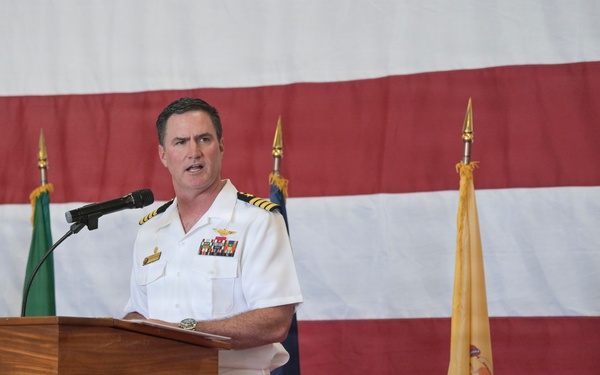 A U.S. Navy officer in white dress uniform is speaking at a podium during a formal military ceremony. The podium bears a seal that reads 'CVN-71' and references 'Theodore Roosevelt,' indicating this is an event associated with the USS Theodore Roosevelt aircraft carrier. Multiple ceremonial flags are displayed in the background alongside a large American flag.