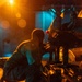 A nighttime scene showing two individuals in dark coveralls/flight suits working on what appears to be military aircraft or heavy equipment on a wet deck surface. The scene is illuminated by warm orange and cool blue artificial lighting, suggesting a naval flight deck or military airfield environment. The workers appear to be performing maintenance or pre-flight checks on the equipment.