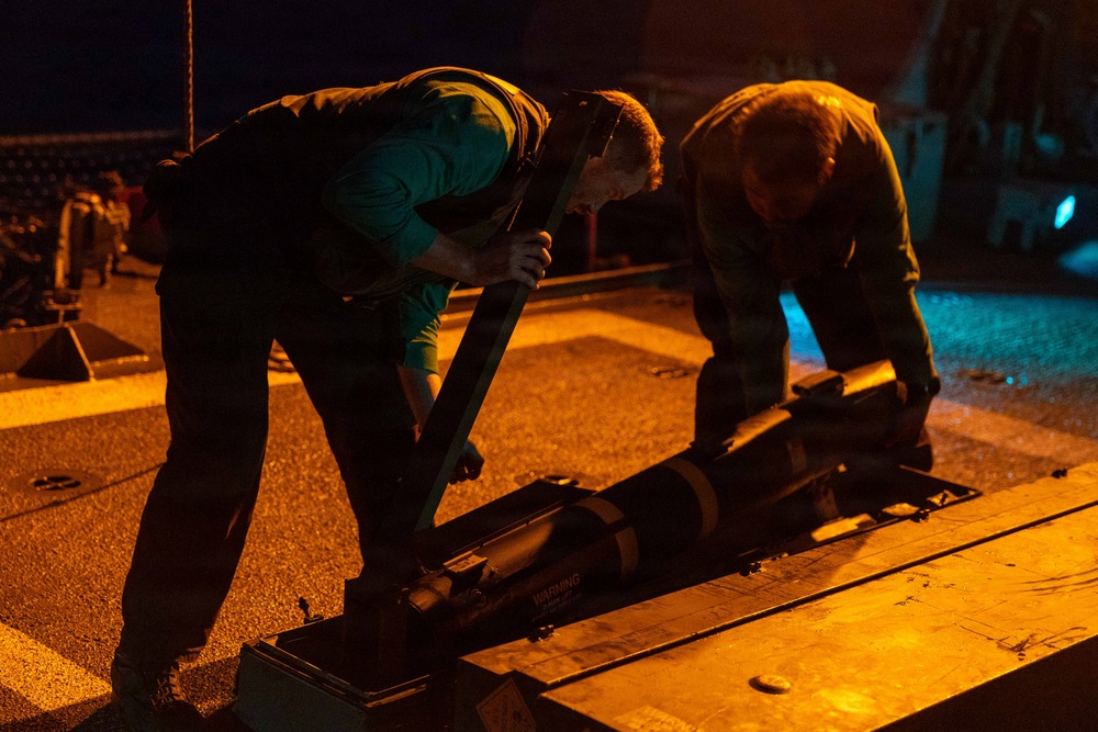 Two individuals are working together at night on what appears to be a military flight deck or similar operational surface, handling a piece of equipment or ordnance on a rail or track system. The scene is illuminated by warm orange/amber lighting, suggesting controlled low-visibility operations. A 'WARNING' label is visible on the equipment being handled.