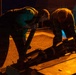 Two individuals are working together at night on what appears to be a military flight deck or similar operational surface, handling a piece of equipment or ordnance on a rail or track system. The scene is illuminated by warm orange/amber lighting, suggesting controlled low-visibility operations. A 'WARNING' label is visible on the equipment being handled.