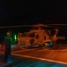 A nighttime naval flight deck operation showing a military helicopter, likely an MH-60 Seahawk, on a ship's flight deck. A deck crew member in the foreground is holding green and red illuminated wands to guide the aircraft. The scene is dramatically lit by orange ambient lighting from the deck, with rotor blur visible from the spinning blades.
