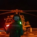 A naval flight deck crew member is marshalling a helicopter on a ship's flight deck during nighttime operations. The handler is using illuminated signal wands — one red and one green — to communicate with the helicopter crew. A second person, likely the helicopter pilot or crew, is visible seated in the aircraft cockpit.