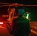 A military flight deck crew member is directing a helicopter during a nighttime flight operations on what appears to be a naval vessel's flight deck. The individual is using illuminated signal wands — one red/orange and one green — to guide the aircraft. The helicopter's rotor blades are visible in motion, captured as light streaks in the long-exposure photograph.