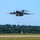 A large military cargo aircraft, consistent with a C-17 Globemaster III, is captured in a low-altitude flight just above a runway, likely during takeoff or a low approach. The aircraft is painted in a dark gray military finish and is viewed from a front-facing angle. The background features a treeline, a water tower, a communications tower, and other distant structures under a clear blue sky.