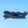 A U.S. Air Force C-17 Globemaster III military cargo aircraft is captured in flight against a clear blue sky. The aircraft bears visible 'U.S. AIR FORCE' markings on the fuselage and 'USAF' on the tail section. The aircraft appears to be climbing or in level cruise flight with landing gear retracted.