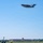 A large military cargo aircraft, consistent with a C-17 Globemaster III, is captured in flight over a military air base on a clear sunny day. The aircraft appears to be in a climbing departure phase with landing gear retracted. Below, a large institutional building with an air traffic control tower is visible on the airfield.