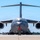 A U.S. Air Force C-17 Globemaster III military cargo aircraft, tail number 0194 assigned to the 445th Airlift Wing, is being marshaled on a tarmac by a ground crew member. The aircraft is photographed head-on, providing a dramatic frontal perspective showing its wide wingspan, four turbofan engines, and T-tail configuration. A marshalller in a red safety vest and headset is directing the aircraft using orange signal wands.