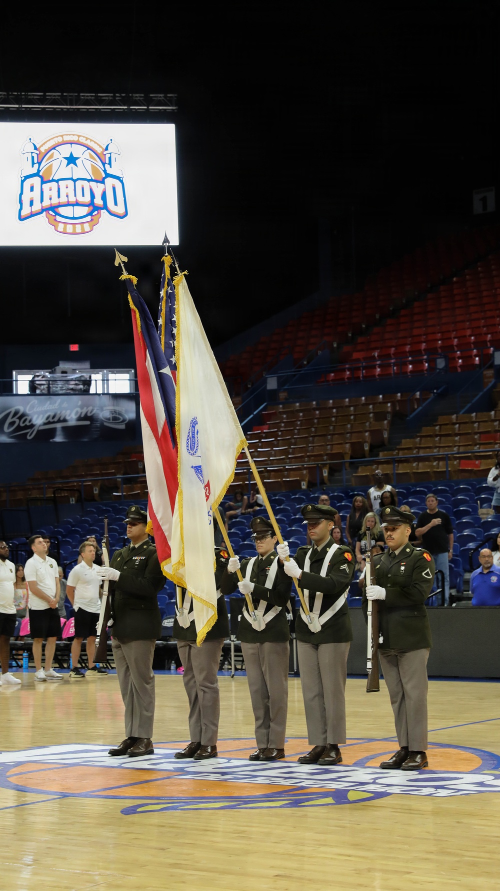 A military color guard ceremony is taking place on a basketball court inside an arena in Bayamón, Puerto Rico. Five soldiers in dress uniforms are presenting flags, including the U.S. flag and what appears to be a unit or organizational flag, at what is identified as the 'Arroyo' basketball classic event. The arena seats are largely empty, suggesting this is a pre-game ceremony.