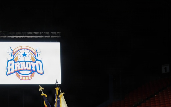A military color guard ceremony is taking place on a basketball court inside an arena in Bayamón, Puerto Rico. Five soldiers in dress uniforms are presenting flags, including the U.S. flag and what appears to be a unit or organizational flag, at what is identified as the 'Arroyo' basketball classic event. The arena seats are largely empty, suggesting this is a pre-game ceremony.