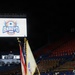 A military color guard ceremony is taking place on a basketball court inside an arena in Bayamón, Puerto Rico. Five soldiers in dress uniforms are presenting flags, including the U.S. flag and what appears to be a unit or organizational flag, at what is identified as the 'Arroyo' basketball classic event. The arena seats are largely empty, suggesting this is a pre-game ceremony.