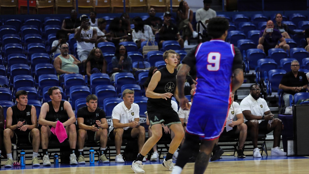 A basketball game is in progress inside an indoor arena with blue seating. One team wears black uniforms and the other wears blue and red uniforms, with player number 9 visible in the foreground. A bench of players and coaching staff in black uniforms is visible along the sideline, with a partially filled audience in the background.