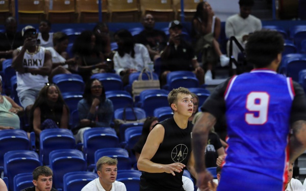 A basketball game is in progress inside an indoor arena with blue seating. One team wears black uniforms and the other wears blue and red uniforms, with player number 9 visible in the foreground. A bench of players and coaching staff in black uniforms is visible along the sideline, with a partially filled audience in the background.