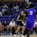 A basketball game is in progress inside an indoor arena with blue seating. One team wears black uniforms and the other wears blue and red uniforms, with player number 9 visible in the foreground. A bench of players and coaching staff in black uniforms is visible along the sideline, with a partially filled audience in the background.