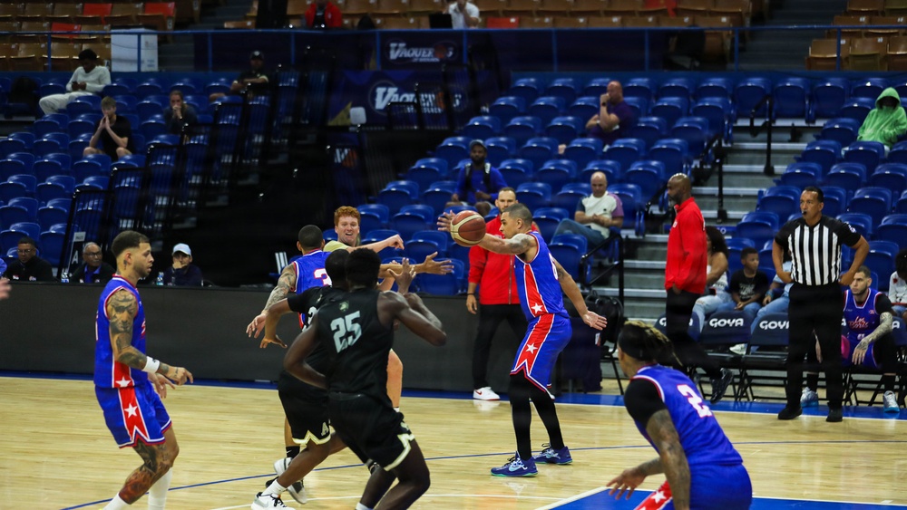 A professional basketball game is in progress inside an indoor arena with blue seating. Two teams are competing — one wearing blue and red uniforms with star logos, and the other wearing black uniforms. The arena appears to have sparse attendance, with most seats empty and only a small number of spectators visible.