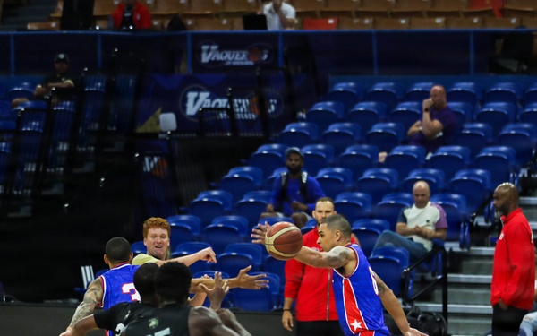 A professional basketball game is in progress inside an indoor arena with blue seating. Two teams are competing — one wearing blue and red uniforms with star logos, and the other wearing black uniforms. The arena appears to have sparse attendance, with most seats empty and only a small number of spectators visible.