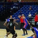 A professional basketball game is in progress inside an indoor arena with blue seating. Two teams are competing — one wearing blue and red uniforms with star logos, and the other wearing black uniforms. The arena appears to have sparse attendance, with most seats empty and only a small number of spectators visible.