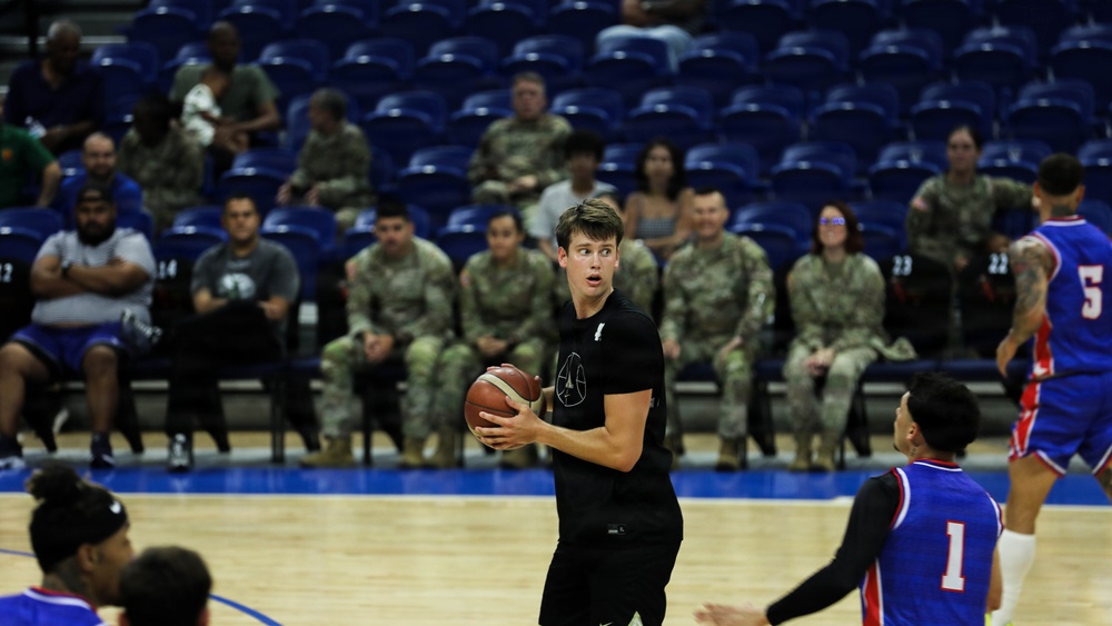 A basketball game is taking place in an indoor arena with blue court flooring and blue stadium seats. A player in a black uniform holds the ball while players in blue and red uniforms defend. A group of military personnel in camouflage uniforms are seated courtside watching the game.