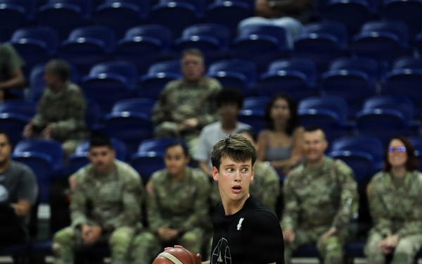 A basketball game is taking place in an indoor arena with blue court flooring and blue stadium seats. A player in a black uniform holds the ball while players in blue and red uniforms defend. A group of military personnel in camouflage uniforms are seated courtside watching the game.