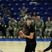 A basketball game is taking place in an indoor arena with blue court flooring and blue stadium seats. A player in a black uniform holds the ball while players in blue and red uniforms defend. A group of military personnel in camouflage uniforms are seated courtside watching the game.