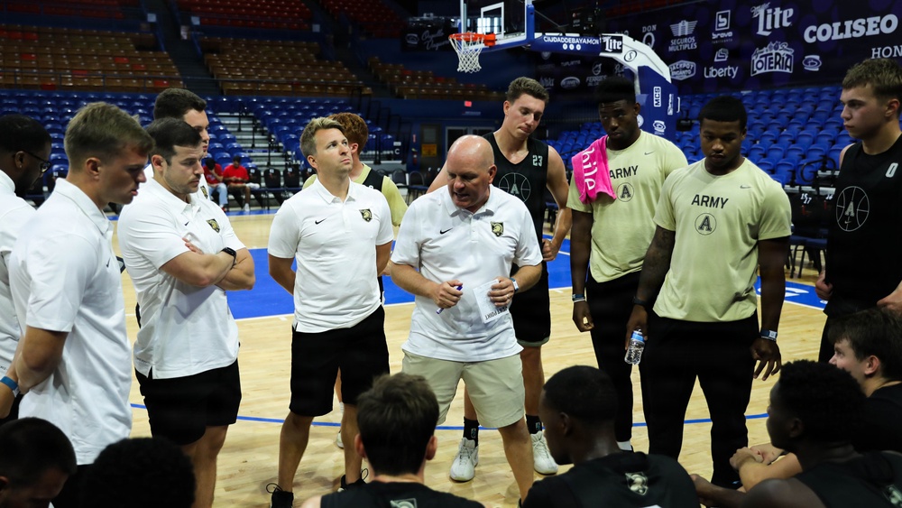 A basketball team huddle is taking place on an indoor court, with a coach in a white Nike polo shirt and white shorts appearing to address the group. Several players are wearing Army-branded athletic wear, suggesting this is the West Point Army Black Knights basketball team. The setting appears to be a professional or semi-professional arena with blue seats visible in the background.