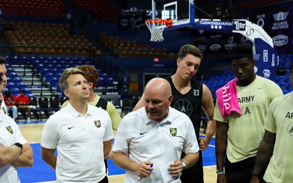 A basketball team huddle is taking place on an indoor court, with a coach in a white Nike polo shirt and white shorts appearing to address the group. Several players are wearing Army-branded athletic wear, suggesting this is the West Point Army Black Knights basketball team. The setting appears to be a professional or semi-professional arena with blue seats visible in the background.