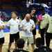 A basketball team huddle is taking place on an indoor court, with a coach in a white Nike polo shirt and white shorts appearing to address the group. Several players are wearing Army-branded athletic wear, suggesting this is the West Point Army Black Knights basketball team. The setting appears to be a professional or semi-professional arena with blue seats visible in the background.