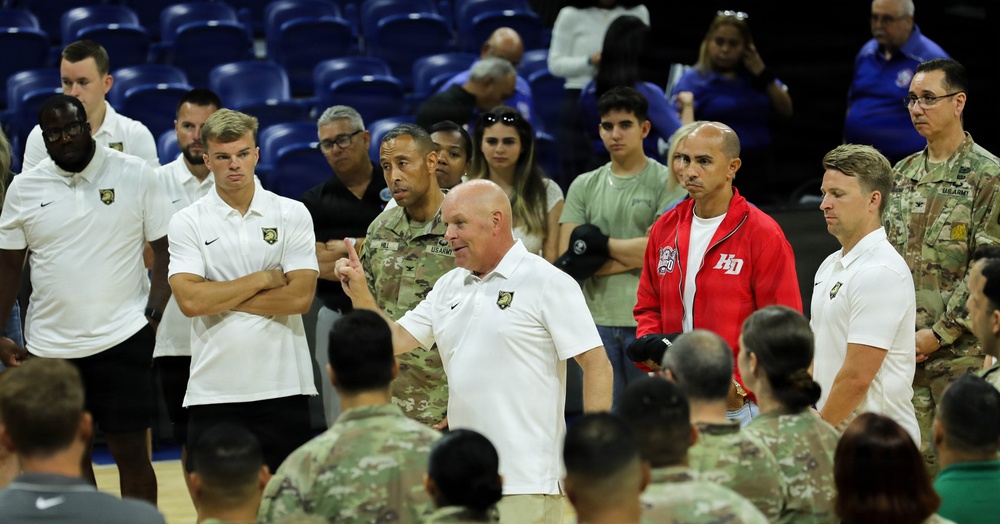 A group of people gathered on what appears to be an indoor basketball court or arena floor. Several individuals are wearing white polo shirts with a military academy logo (West Point/Army), while others are in U.S. Army camouflage uniforms. A man in a white polo shirt appears to be addressing the group, suggesting a team or organizational meeting.