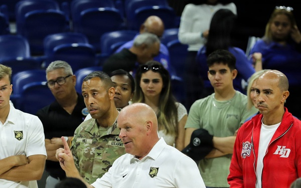 A group of people gathered on what appears to be an indoor basketball court or arena floor. Several individuals are wearing white polo shirts with a military academy logo (West Point/Army), while others are in U.S. Army camouflage uniforms. A man in a white polo shirt appears to be addressing the group, suggesting a team or organizational meeting.