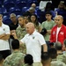 A group of people gathered on what appears to be an indoor basketball court or arena floor. Several individuals are wearing white polo shirts with a military academy logo (West Point/Army), while others are in U.S. Army camouflage uniforms. A man in a white polo shirt appears to be addressing the group, suggesting a team or organizational meeting.