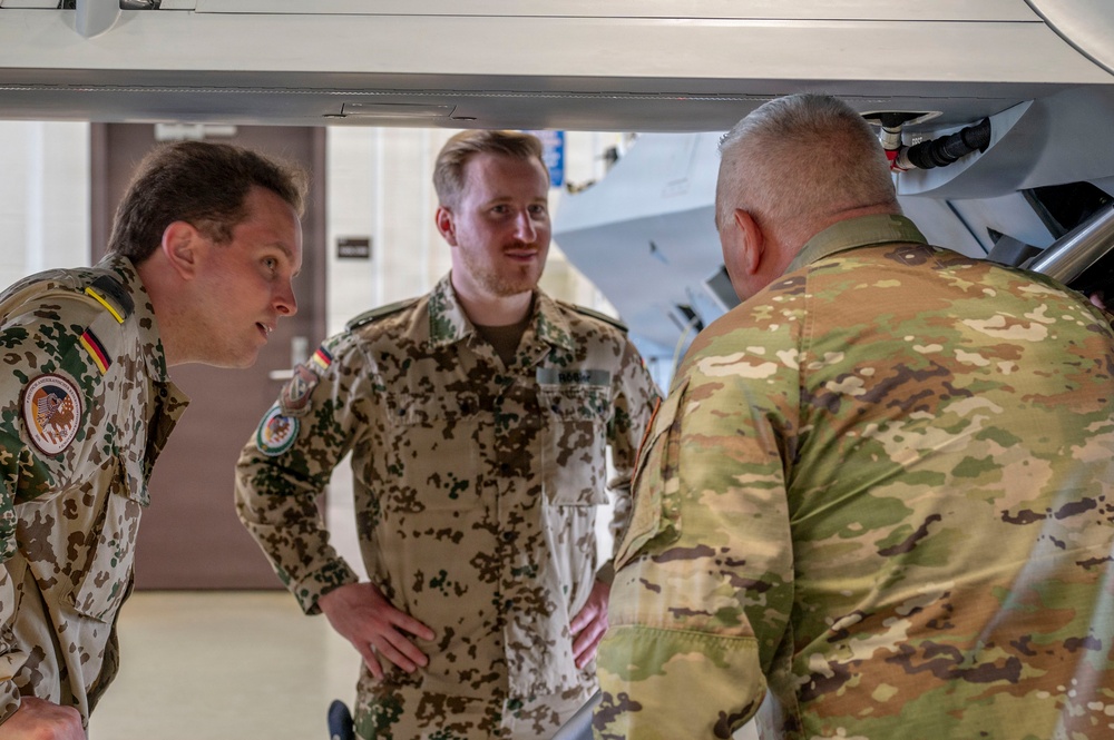 Three military personnel are engaged in a discussion in what appears to be an indoor facility, possibly a hangar or exhibition space. Two individuals are wearing German Bundeswehr flecktarn camouflage uniforms with visible German flag patches, while the third wears a US Army OCP (Operational Camouflage Pattern) uniform. They appear to be examining or discussing equipment visible in the foreground and background.