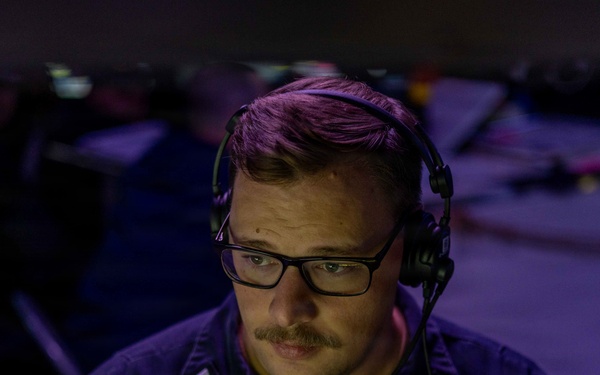 A military officer, identified by a name tag partially reading 'CARTHY', is seated at a workstation in a dimly lit operations center illuminated by purple/blue ambient light. He is wearing a dark naval uniform with rank insignia and a communications headset, focused intently on monitors in front of him. The setting appears to be a combat information center or similar tactical operations environment aboard a naval vessel.
