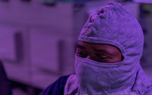 A person wearing a gray fabric hood/balaclava that covers most of their face is visible in a purple-tinted environment. Only their eyes are exposed through the hood opening. The background suggests an industrial or technical setting with equipment visible.