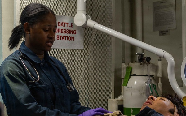 A military medical corpsman wearing a stethoscope and purple gloves is treating a patient lying on a stretcher at a Battle Dressing Station aboard what appears to be a naval vessel. The patient is lying supine on an orange rescue/litter stretcher and appears to have a tourniquet applied to their leg. At least two other personnel are partially visible assisting with the treatment.