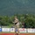 A military service member in OCP (Operational Camouflage Pattern) uniform is throwing a ceremonial first pitch on a baseball field. The field features red clay mound and green artificial turf, with outfield advertising banners visible. The backdrop includes a forested mountain, suggesting a location in Alaska based on the 'Go Chinooks' banner and mountainous terrain.