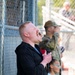 A red-haired man in a black jacket is singing passionately into a handheld microphone, likely performing the national anthem or a similar patriotic song at an outdoor event. Behind him stand several individuals including a person in military camouflage uniform and a young girl in a light blue shirt, both with hands over their hearts. The setting appears to be a sports field or outdoor venue with chain-link fencing visible in the background.