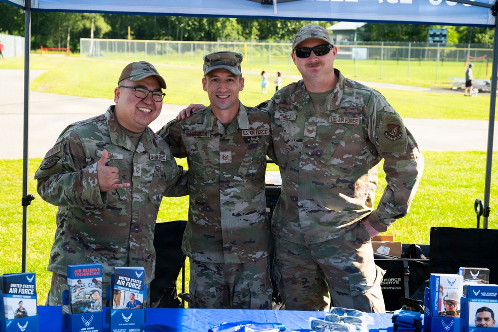 Three U.S. Air Force personnel in OCP (Operational Camouflage Pattern) uniforms pose together at an outdoor recruitment or outreach booth. The table in front of them displays Air Force Reserve and U.S. Air Force recruitment brochures and materials. The event appears to be taking place at an outdoor venue with a green field visible in the background.